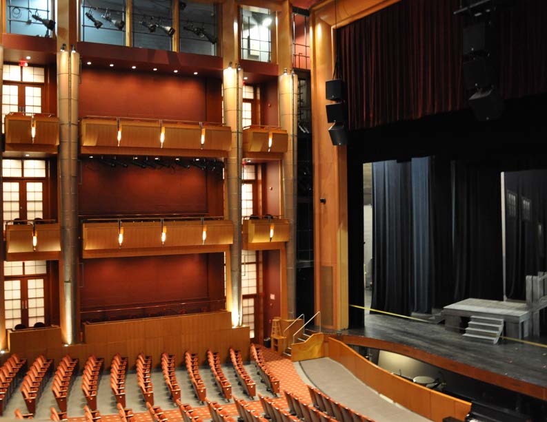 Interior of a modern theater with tiered balconies, rows of seats, and a stage with curtains.
